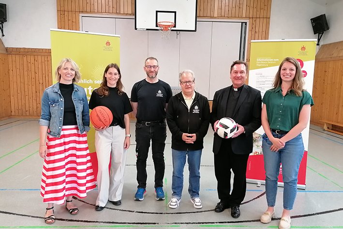 Sechs Personen, darunter ein Priester, halten einen Basketball und Fußball in einer Turnhalle, mit Don Bosco Augsburg Bannern.