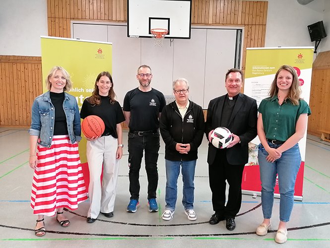 Sechs Personen, darunter ein Priester, halten einen Basketball und Fußball in einer Turnhalle, mit Don Bosco Augsburg Bannern.