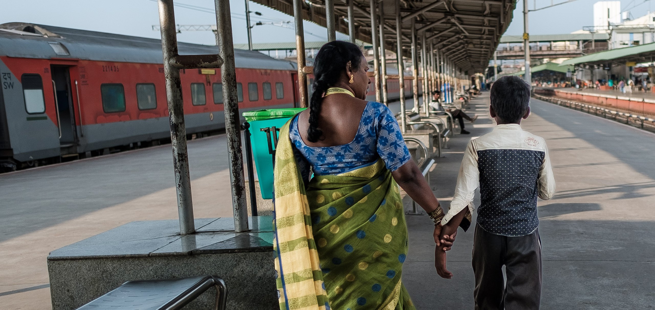 Eine indische Frau im Sari hält die Hand eines Jungen, sie gehen über einen Bahnsteig mit einem wartenden Zug.