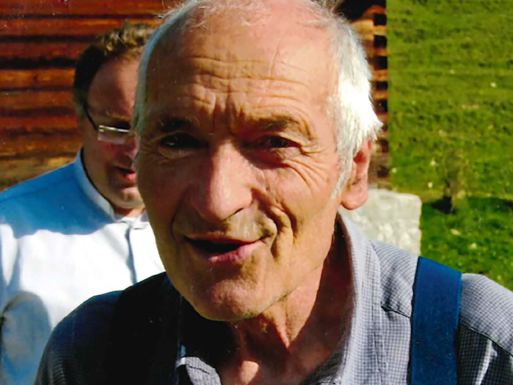 Nahaufnahme eines älteren Mannes mit weißen Haaren und blauen Hosenträgern, lächelnd im Freien. Ein weiterer Mann ist unscharf im Hintergrund.