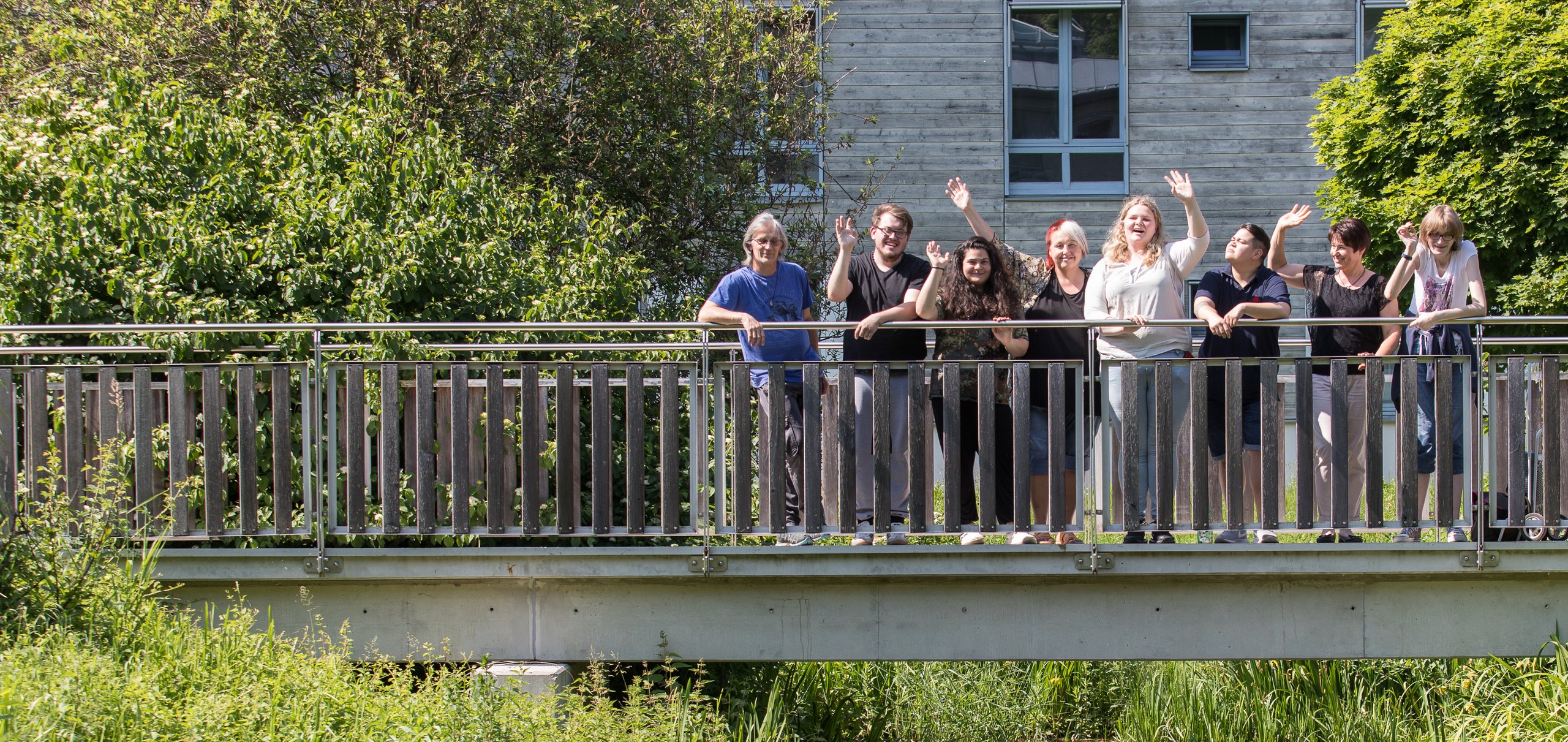 Neun Personen winken lächelnd von einer Brücke, umgeben von üppigem Grün und einem Gebäude im Hintergrund.