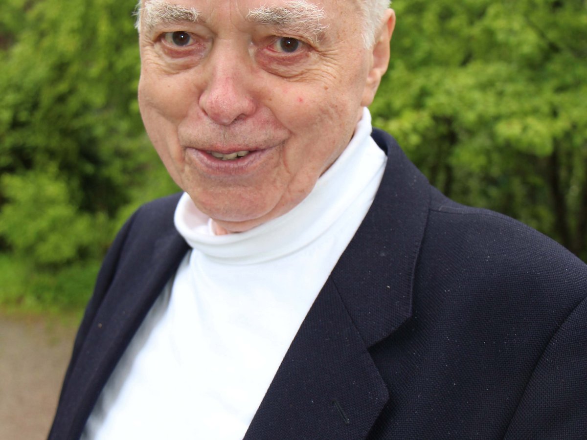 Ein lächelnder älterer Mann mit weißen Haaren, der einen weißen Rollkragenpullover und eine dunkelblaue Jacke trägt, vor grünem Hintergrund.