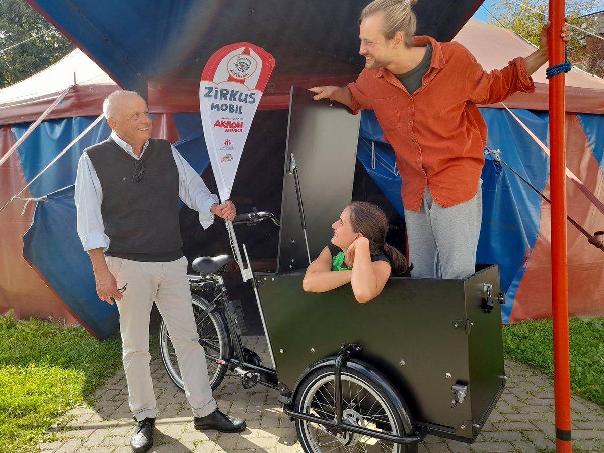 Drei lächelnde Personen mit einem Zirkus Mobil Lastenrad und einem bunten Zirkuszelt im Hintergrund.