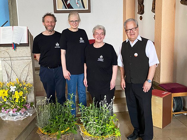 Vier Personen, drei in schwarzen "Don Bosco Mission" T-Shirts und einer in einer Weste, lächeln in einer Kirche mit Blumen.