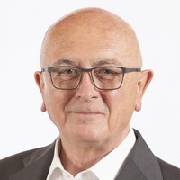 Porträt eines glatzköpfigen älteren Mannes mit Brille und dunklem Anzug, leicht lächelnd vor weißem Hintergrund.