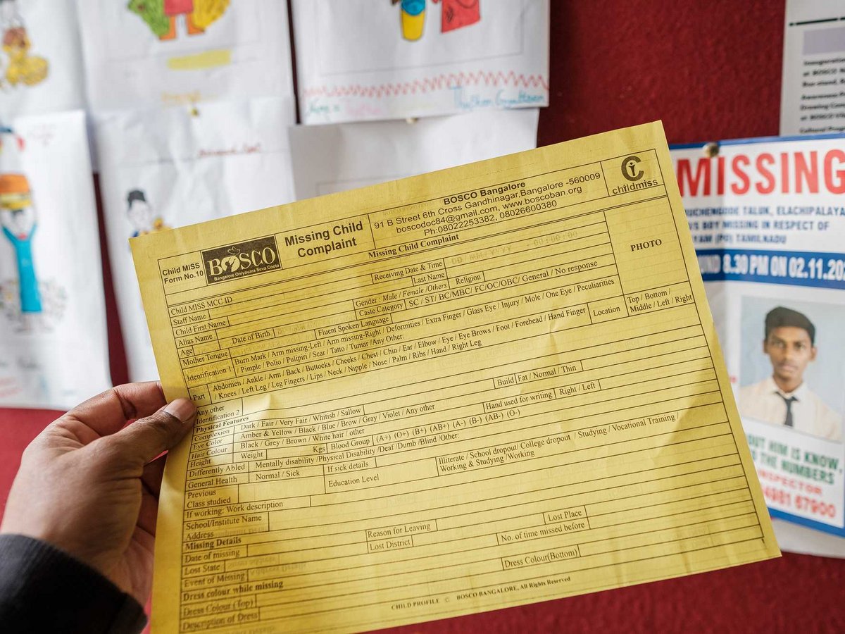 Eine Hand hält ein gelbes Formular für eine Vermisstenanzeige von BOSCO Bangalore, im Hintergrund ein Poster.