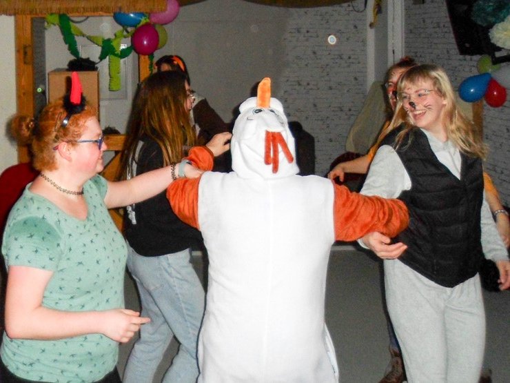 Vier Personen tanzen im Kreis auf einer Party, eine im Schneemannkostüm, eine mit Teufelshörnern, eine mit Katzengesicht.