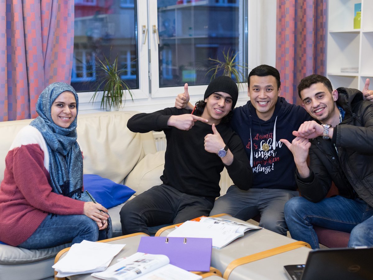 Vier lächelnde junge Erwachsene, darunter eine Frau mit Hidschab, sitzen auf einer Couch und lernen. Sie zeigen Daumen hoch.