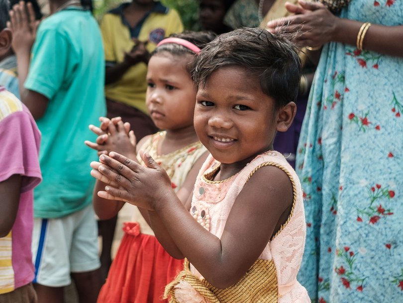 Eine Gruppe lächelnder Kinder und Erwachsener, einige mit zusammengelegten Händen zur Begrüßung, steht draußen unter einem einfachen Unterstand.