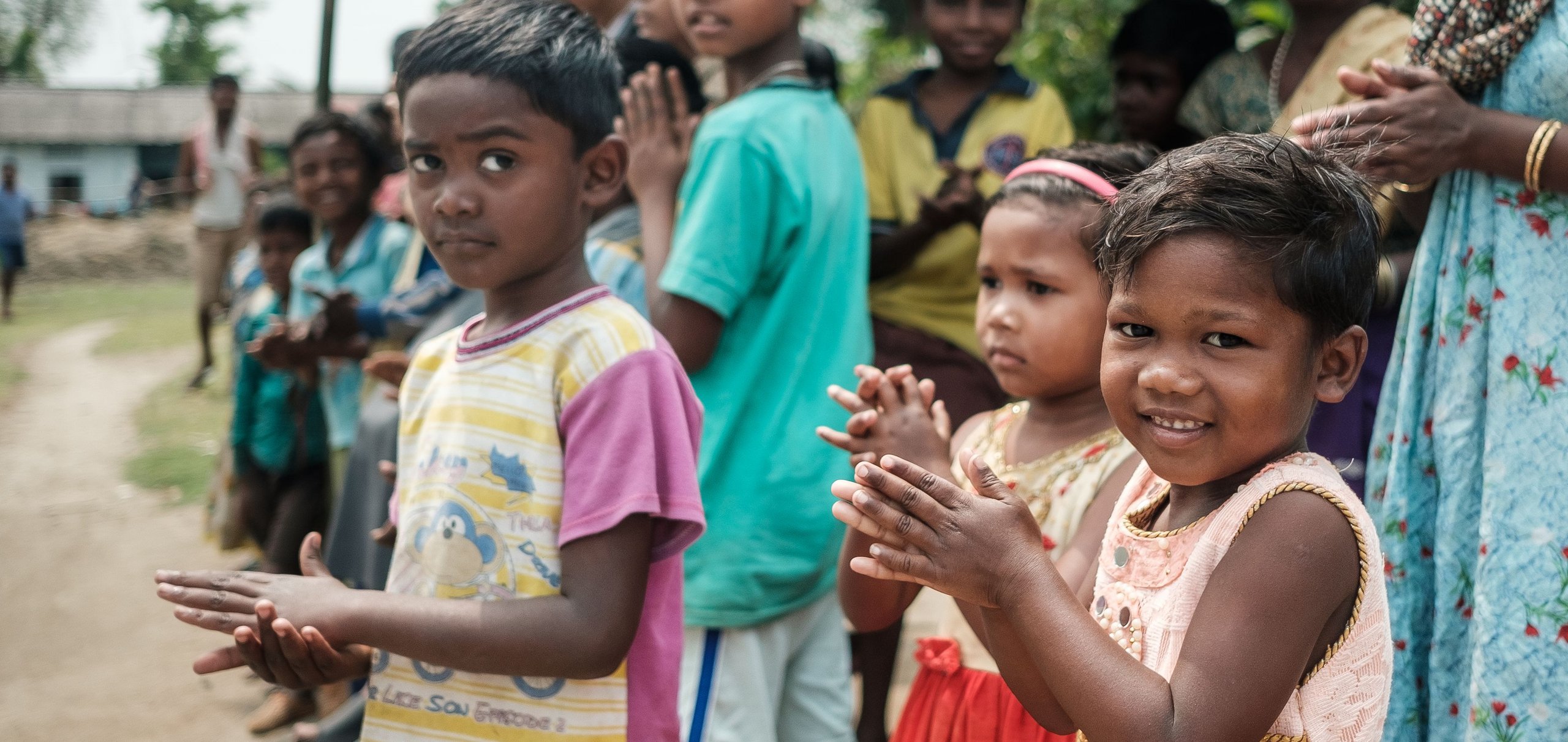 Eine Gruppe lächelnder Kinder und Erwachsener, einige mit zusammengelegten Händen zur Begrüßung, steht draußen unter einem einfachen Unterstand.