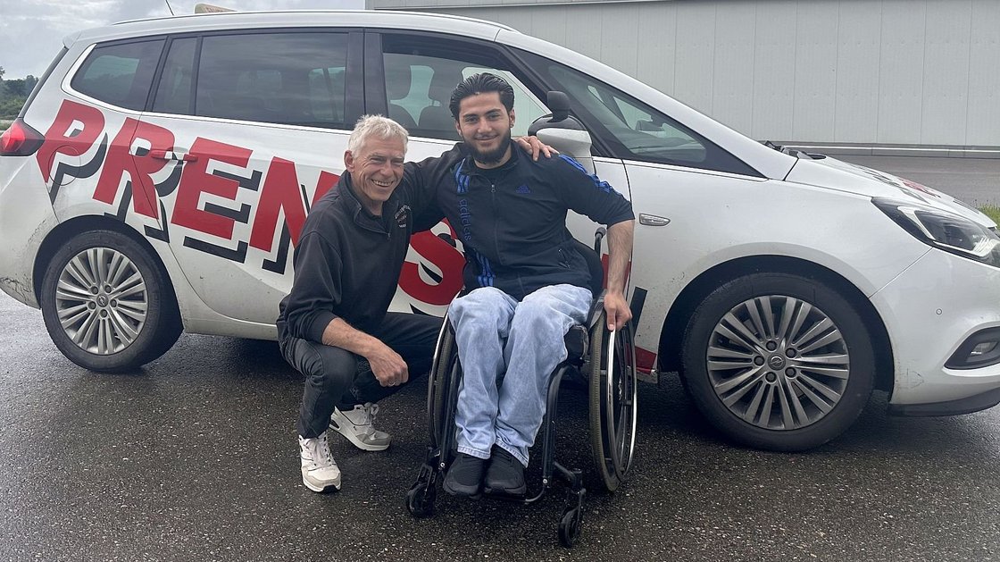 Ein Fahrlehrer und ein Fahrschüler im Rollstuhl stehen vor einem Fahrschulauto.