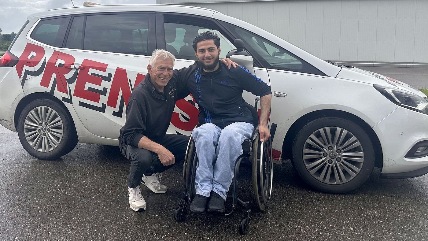 Ein Fahrlehrer und ein Fahrschüler im Rollstuhl stehen vor einem Fahrschulauto.