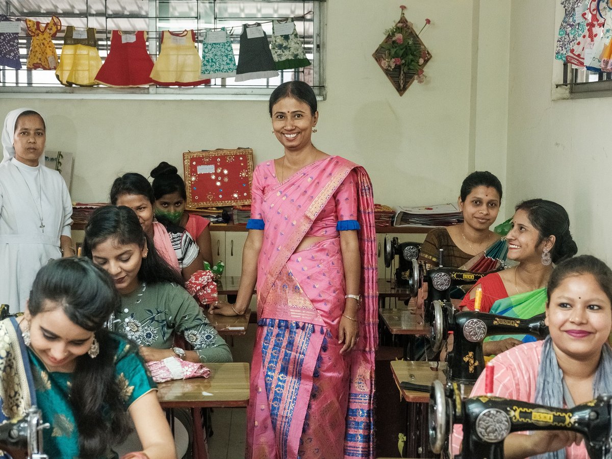 Frauen und eine Nonne lächeln in einer Nähwerkstatt, einige bedienen Nähmaschinen, bunte Kleider hängen darüber.