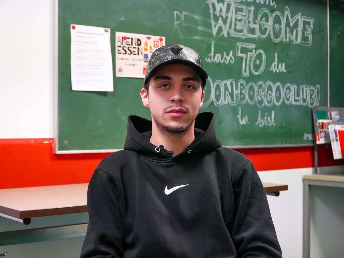 Junger Mann in schwarzem Hoodie und gemusterter Cap blickt in die Kamera, im Hintergrund eine grüne Tafel.