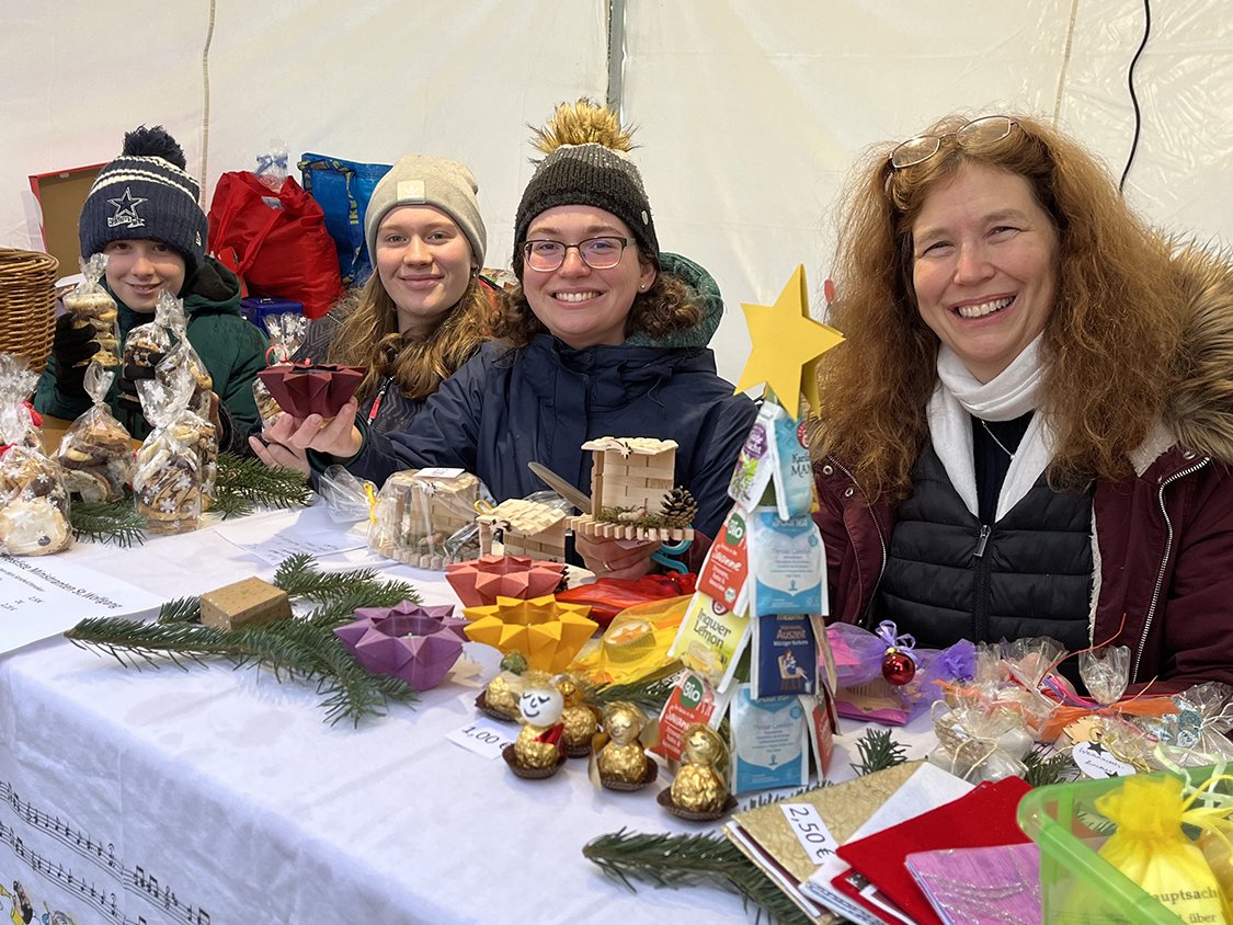 Vier lächelnde Personen, darunter ein Junge und drei Frauen, hinter einem Marktstand mit Gebäck und Handwerk.
