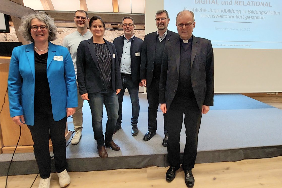 Sechs Personen, darunter ein Geistlicher, stehen lächelnd auf einer Bühne vor einem Bildschirm über kirchliche Jugendbildung.