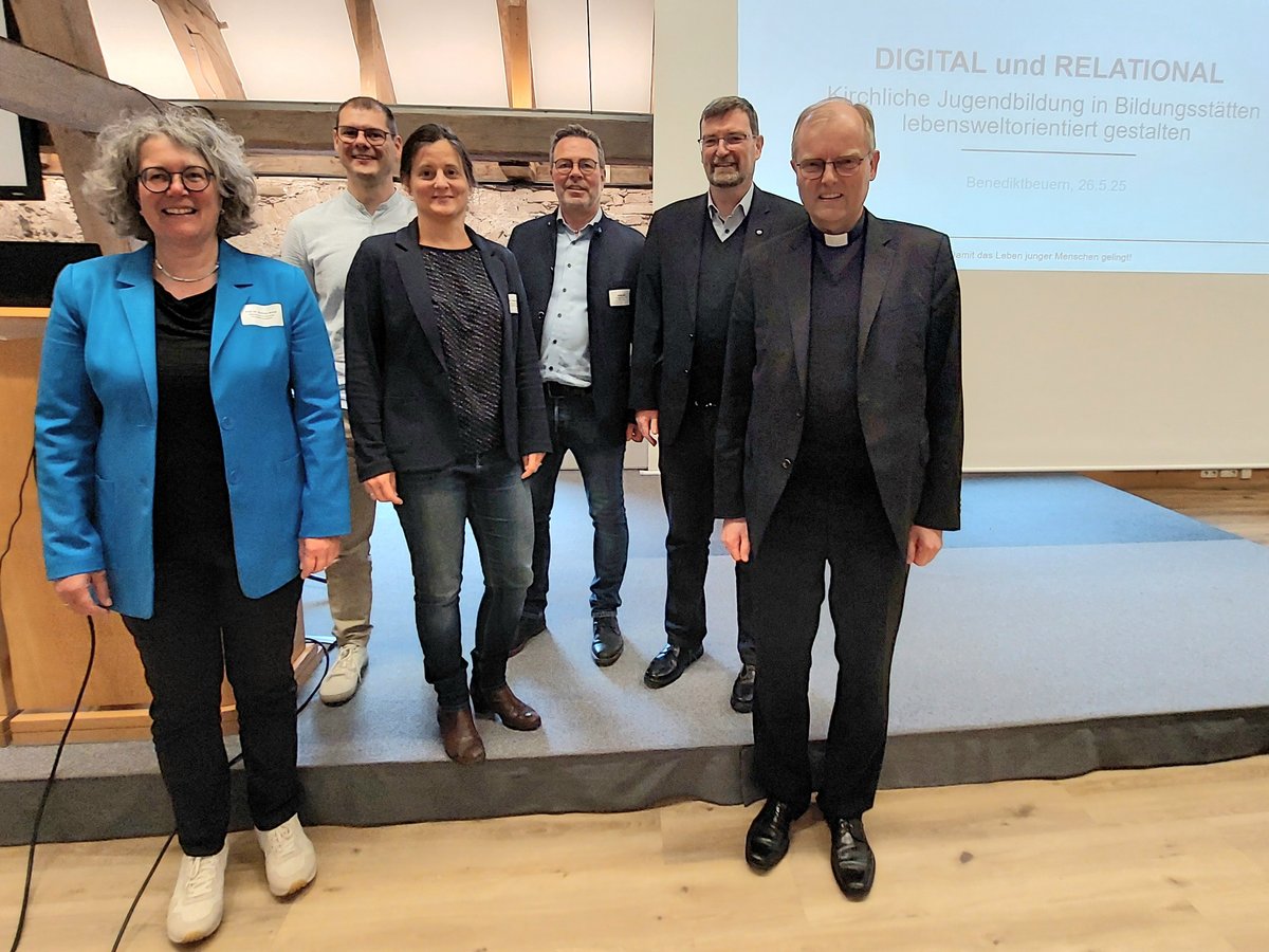 Sechs Personen, darunter ein Geistlicher, stehen lächelnd auf einer Bühne vor einem Bildschirm über kirchliche Jugendbildung.