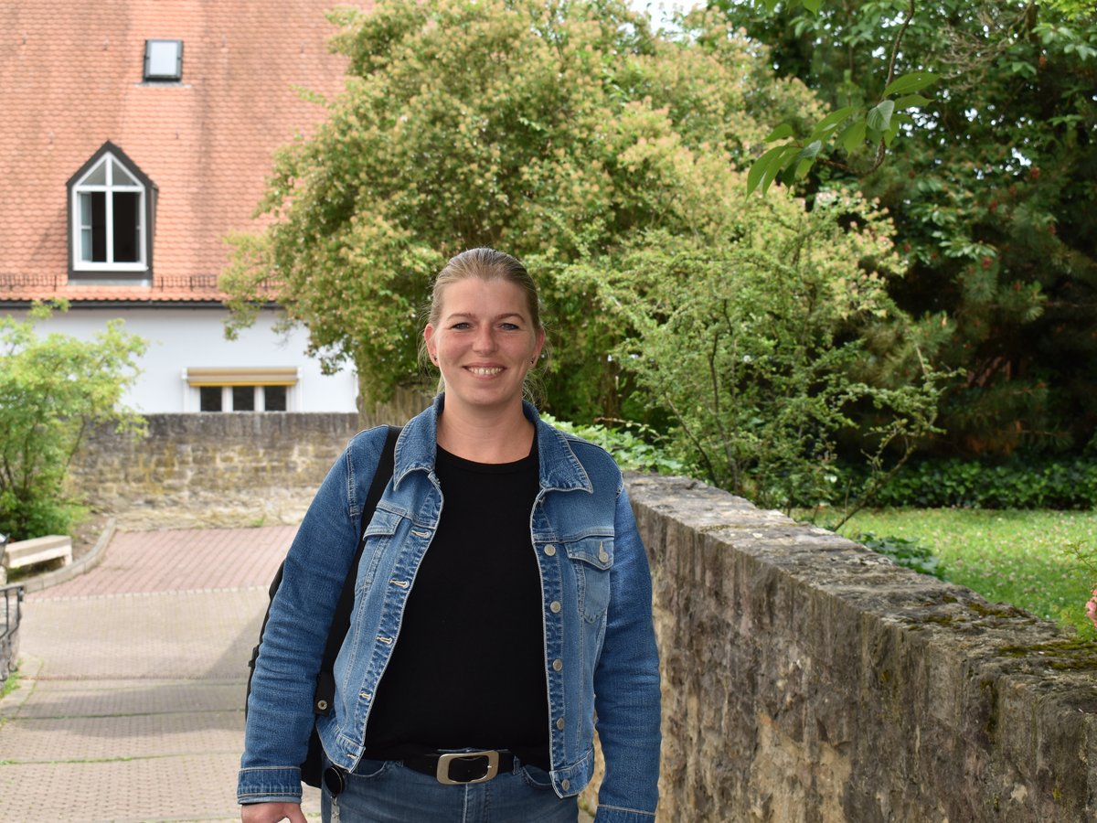 Lachende Frau in Jeansjacke vor Steinmauer, Bäumen und einem alten Gebäude mit Ziegeldach.
