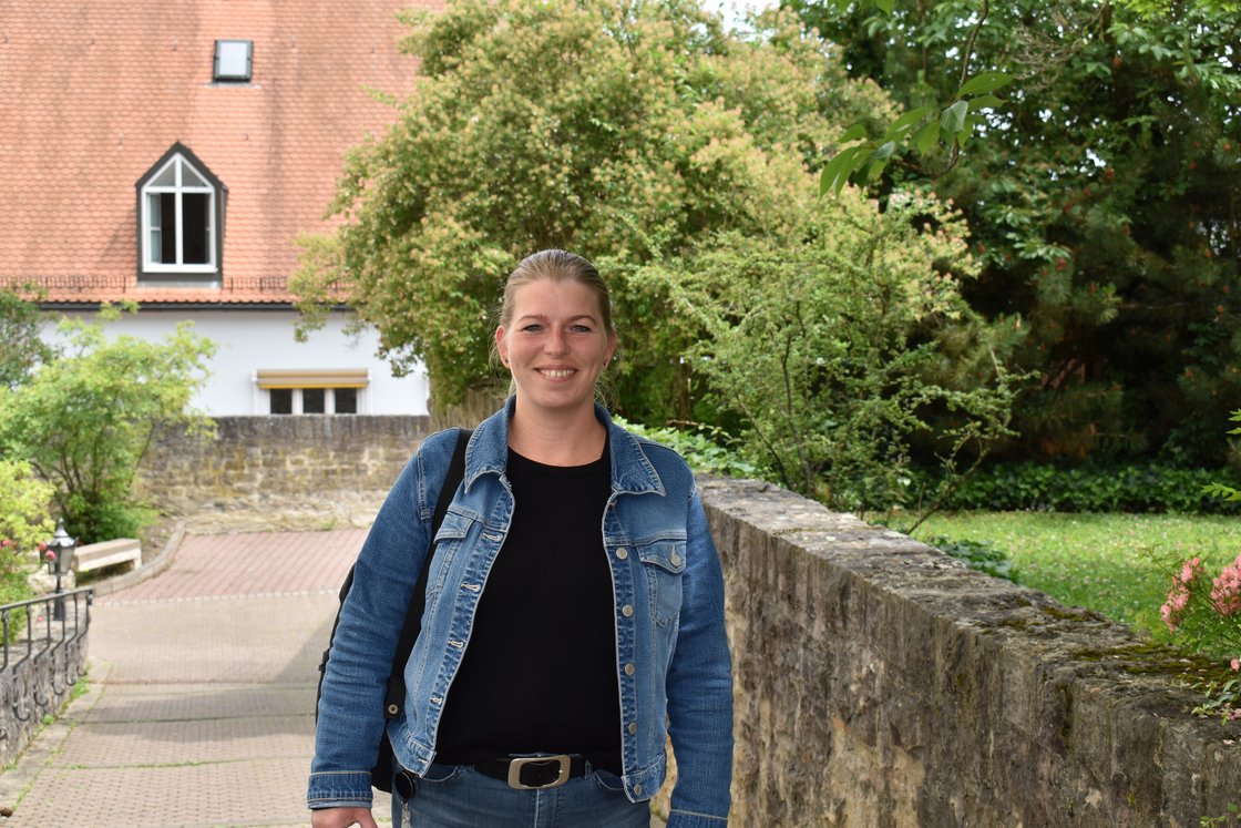 Lachende Frau in Jeansjacke vor Steinmauer, Bäumen und einem alten Gebäude mit Ziegeldach.