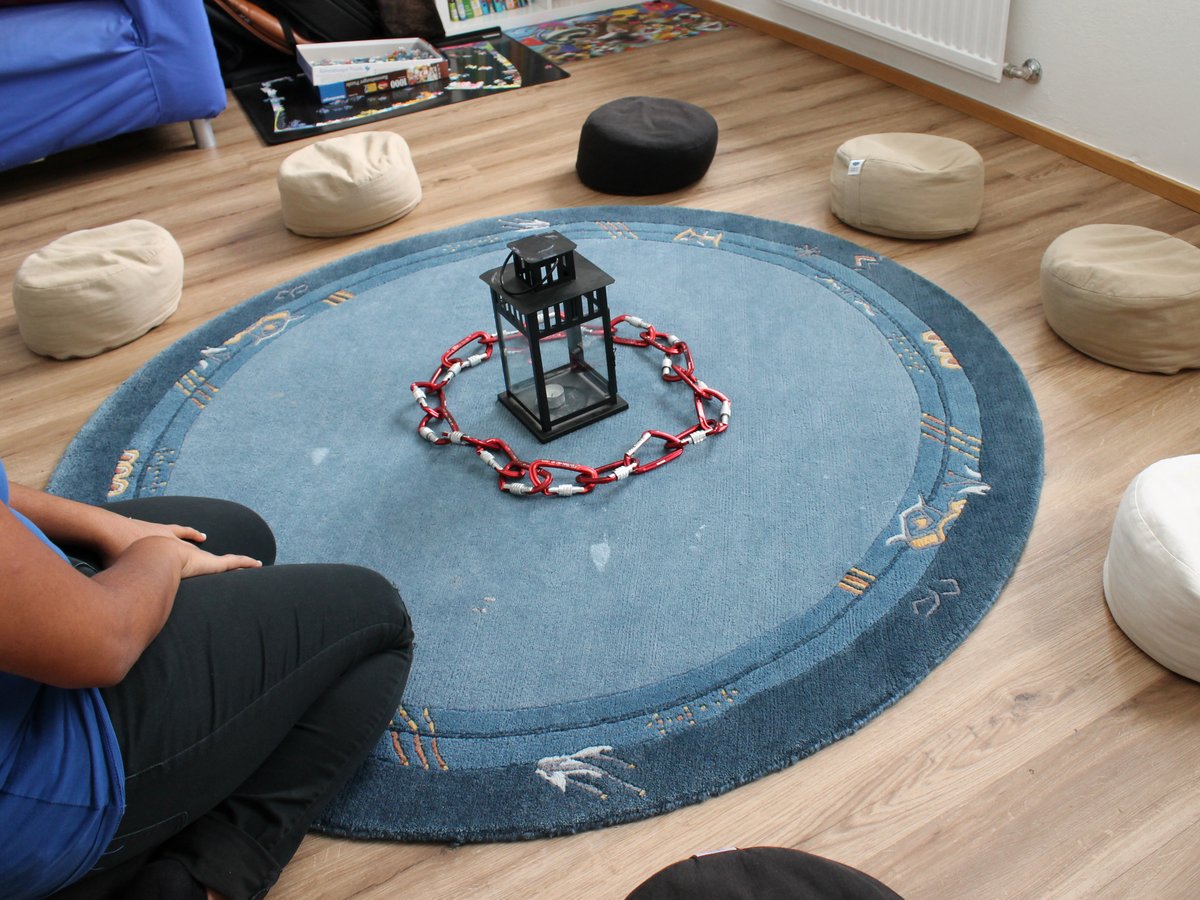 Ein runder blauer Teppich mit Laterne und roter Karabinerkette in der Mitte, umgeben von Sitzkissen; eine Person sitzt daneben.