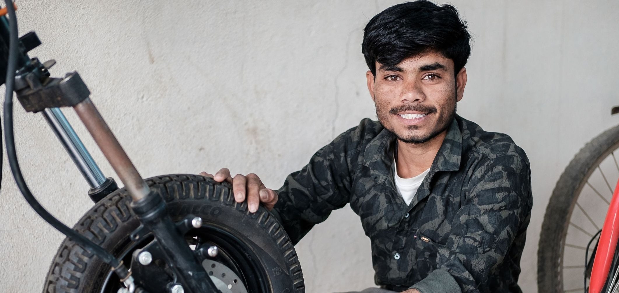 Ein lächelnder Mann in Tarnkleidung arbeitet mit einem Schraubenschlüssel am Vorderrad eines Rollers.