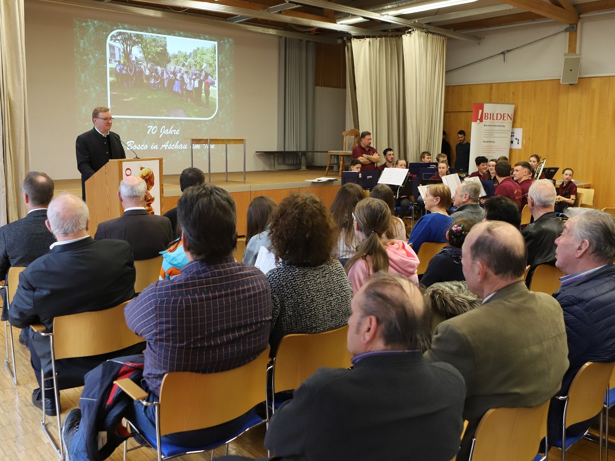 Ein Mann hält eine Rede vor einem Publikum, während eine Projektion '70 Jahre Bosco' zeigt und eine Jugendband spielt.