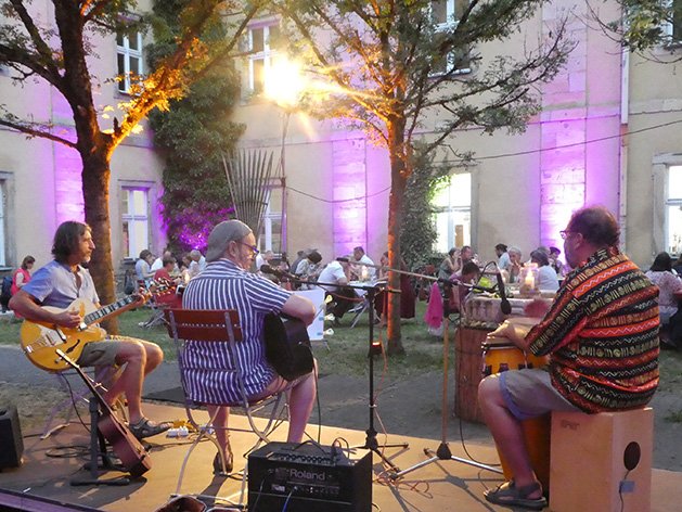 Open-Air-Konzert mit drei Musikern, die Gitarre und Schlagzeug auf einer Bühne spielen, mit Menschen und beleuchteten Gebäuden im Hintergrund.