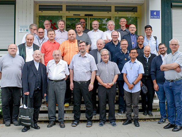 Große Gruppe von Männern, viele in geistlicher Kleidung, posiert für ein Gruppenfoto im Freien vor einem Gebäude mit Straßenschild.