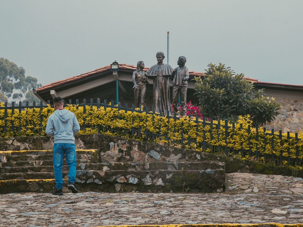 Rückansicht eines Mannes auf einem Steinweg, der eine Bronzestatue von drei Figuren hinter einem Zaun betrachtet.