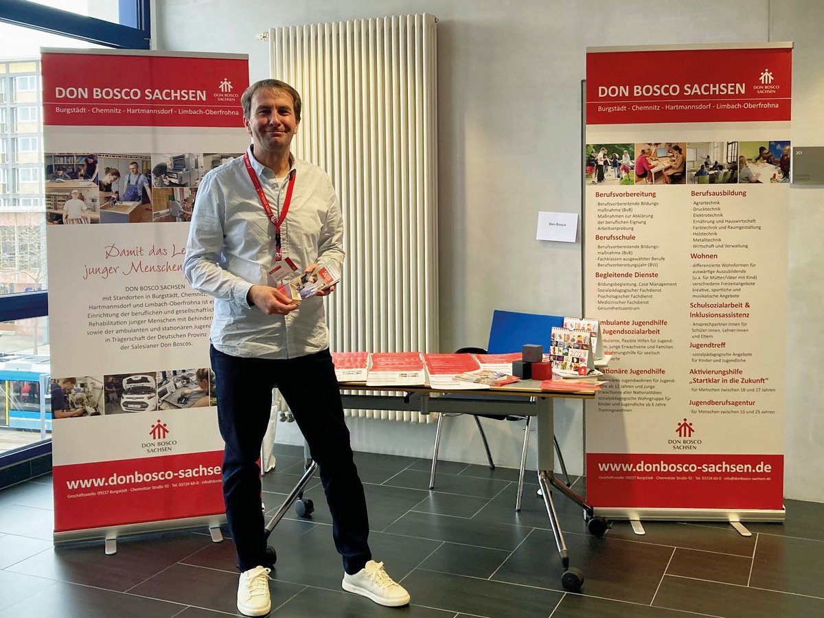 Lächelnder Mann an einem Don Bosco Sachsen Infostand, Broschüren haltend, mit Dienstleistungsbannern dahinter.