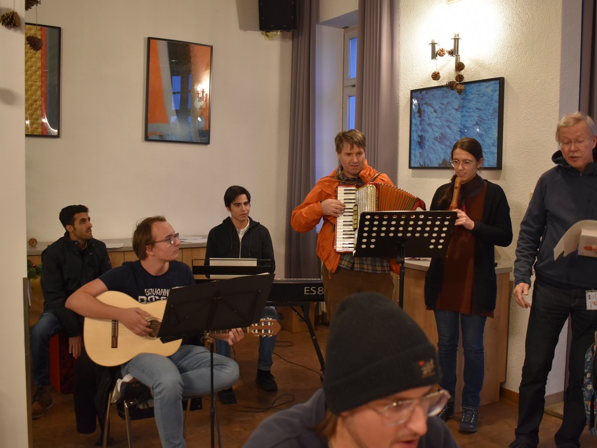 Eine Gruppe von Menschen spielt Musikinstrumente wie Akkordeon, Gitarre und Blockflöte in einem gemütlichen Raum.