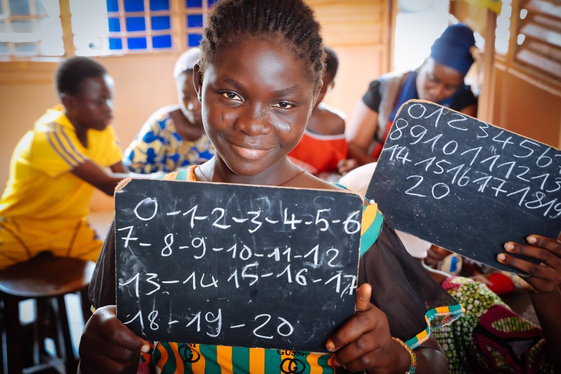Lächelndes afrikanisches Mädchen hält eine Tafel mit Zahlen 0-20, umgeben von anderen Kindern in einem Klassenzimmer.