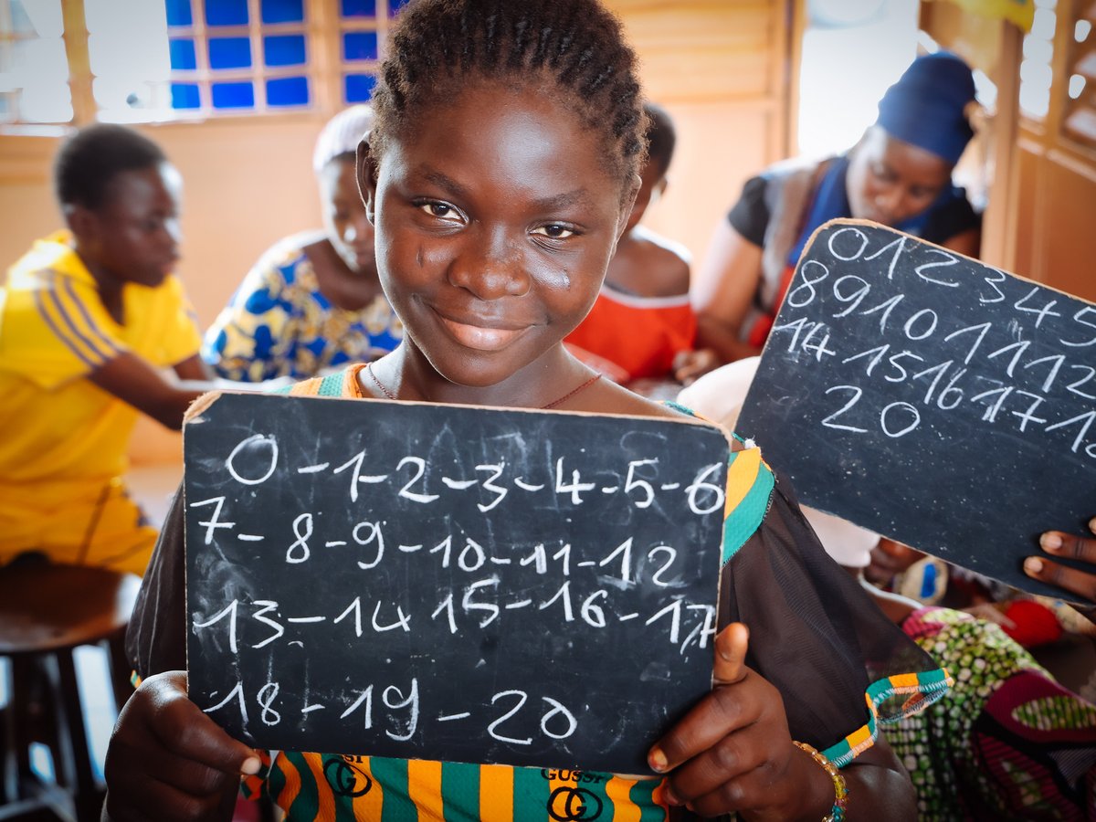 Lächelndes afrikanisches Mädchen hält eine Tafel mit Zahlen 0-20, umgeben von anderen Kindern in einem Klassenzimmer.