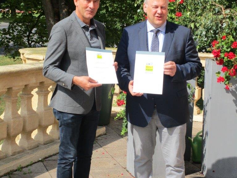 Zwei lächelnde Männer im Freien halten Dokumente, möglicherweise im Zusammenhang mit einem Umweltpakt.