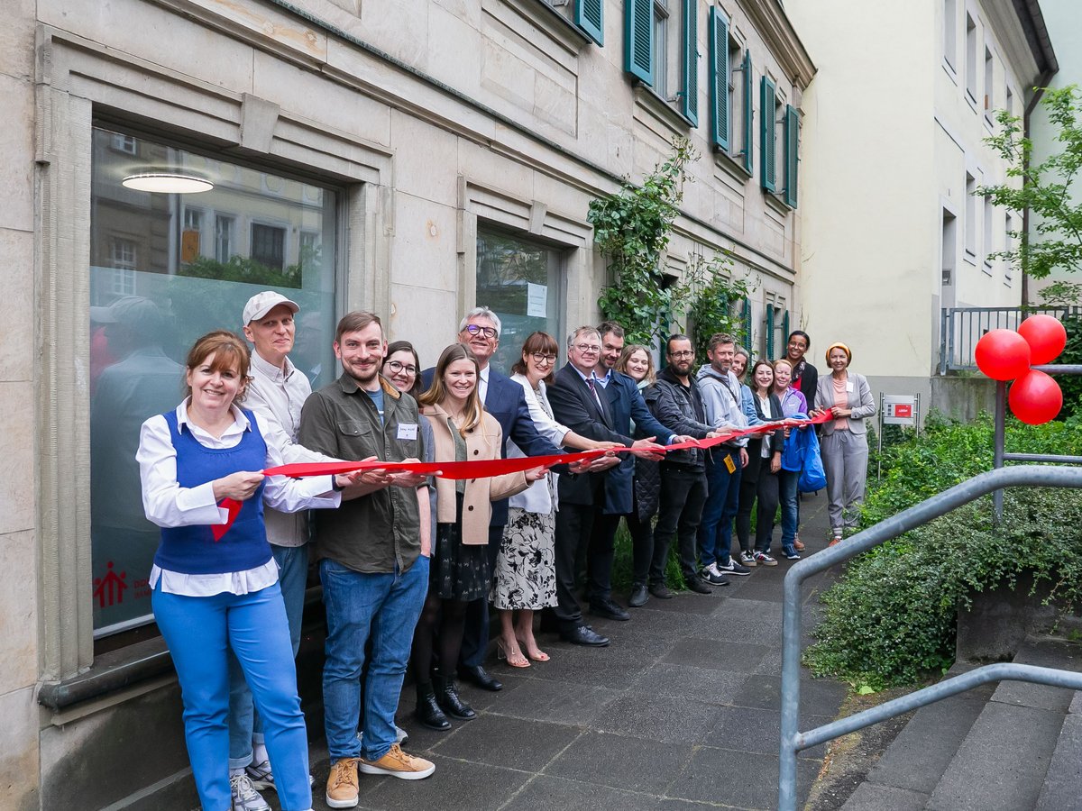 Eine Gruppe lächelnder Menschen schneidet ein rotes Band bei einer Eröffnungszeremonie vor einem Gebäude mit roten Ballons.