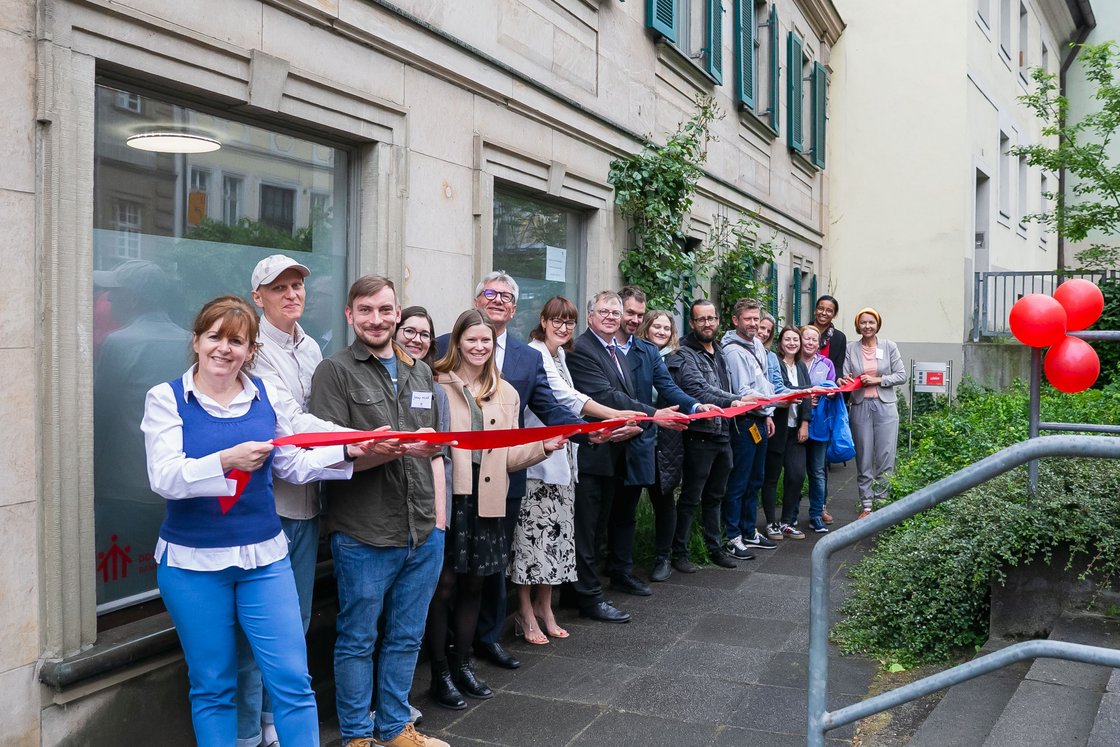Eine Gruppe lächelnder Menschen schneidet ein rotes Band bei einer Eröffnungszeremonie vor einem Gebäude mit roten Ballons.