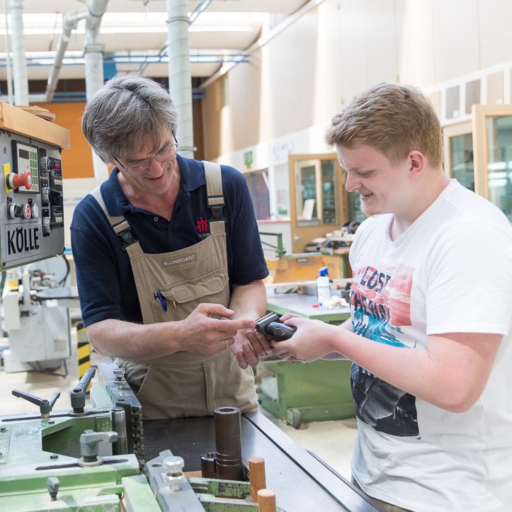 Ein Ausbilder in Arbeitskleidung zeigt einem lächelnden jungen Mann ein kleines Gerät in einer Holzwerkstatt.