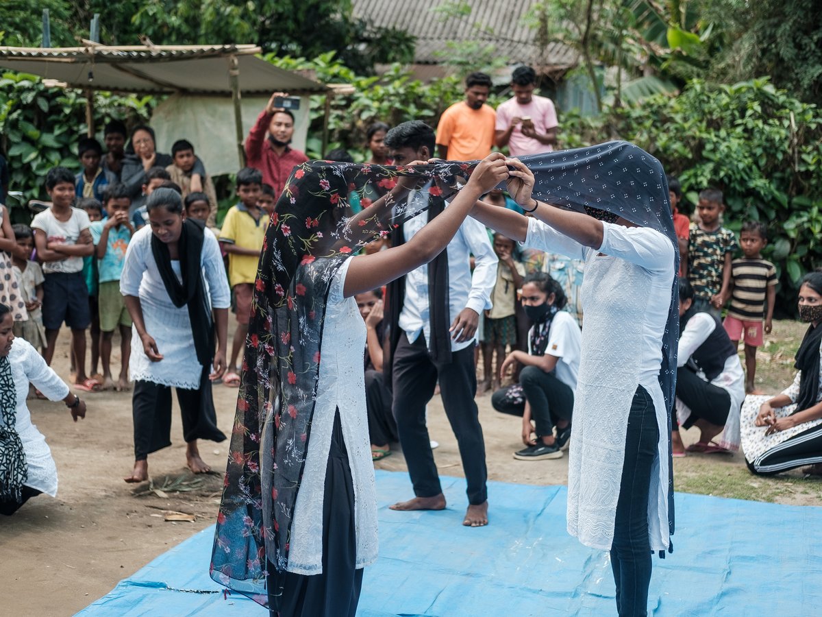 Zwei Frauen halten einen gemusterten Stoff hoch, beobachtet von Kindern und Erwachsenen im Freien.