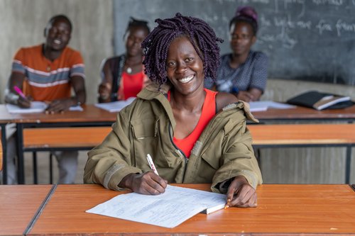 Eine lächelnde junge Frau mit lila Dreadlocks schreibt in einem Klassenzimmer, andere Schüler und eine Tafel im Hintergrund.