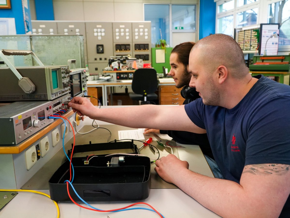 Zwei Männer in einem Elektroniklabor. Einer justiert ein Messgerät, dessen Kabel zu einem offenen Gehäuse mit Motor führen.