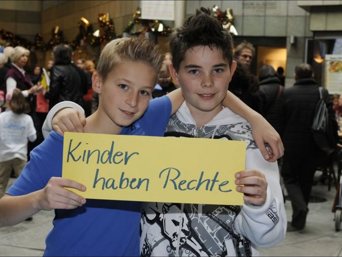 Zwei lächelnde Jungen halten ein gelbes Schild mit der Aufschrift 'Kinder haben Rechte' in einem belebten Innenraum.