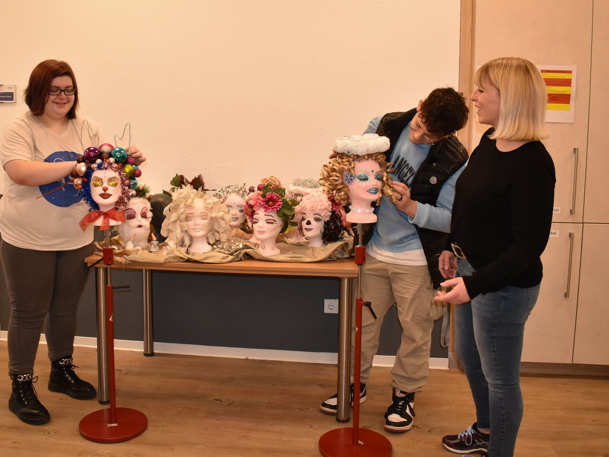Zwei Frauen und ein Mann schmücken Schaufensterpuppenköpfe kreativ mit festlichen und fantasievollen Accessoires.