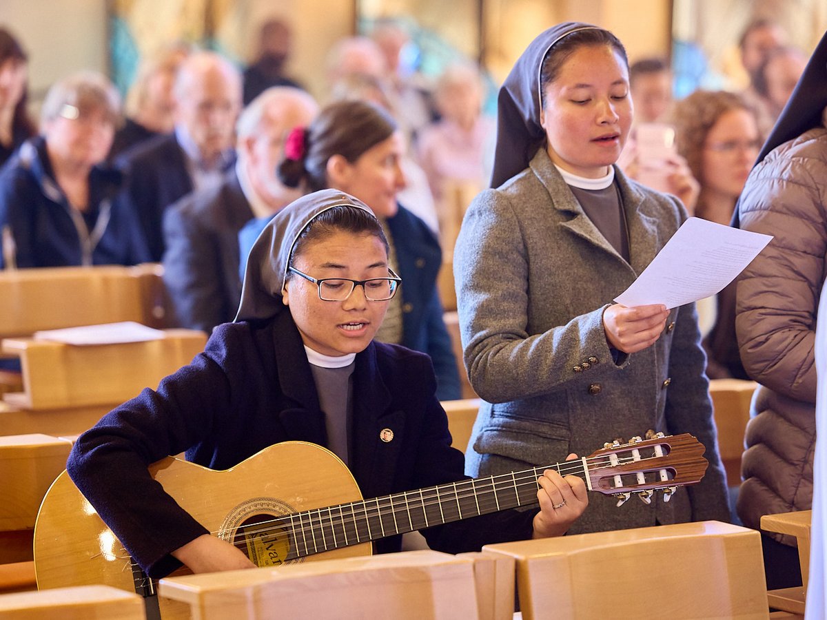 Zwei Nonnen singen und musizieren Gitarre in einem Gottesdienst. Eine hält ein Notenblatt, die andere spielt Gitarre.
