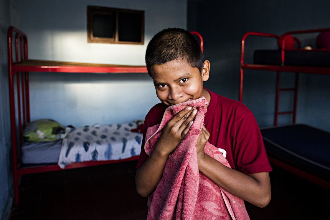 Junger Junge in rotem Shirt lächelt schüchtern, hält ein rosa Handtuch vor den Mund, in blauem Raum mit Etagenbetten.