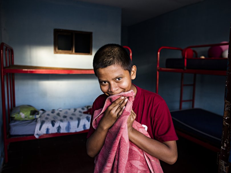 Junger Junge in rotem Shirt lächelt schüchtern, hält ein rosa Handtuch vor den Mund, in blauem Raum mit Etagenbetten.