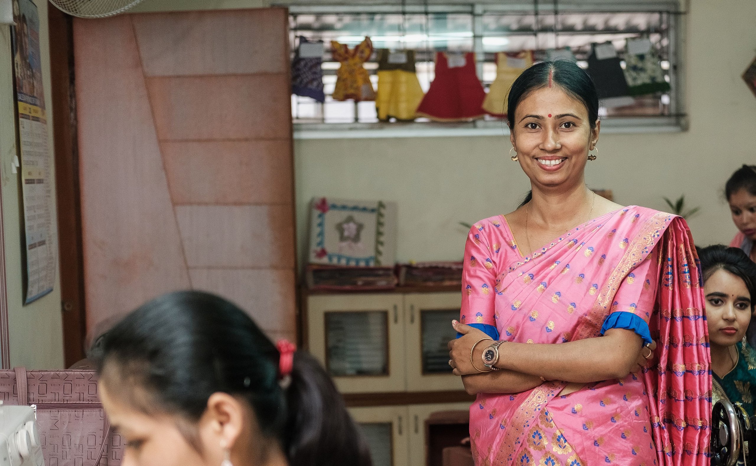 Lächelnde Frau im pinkfarbenen Sari in einer geschäftigen Nähwerkstatt, umgeben von anderen Frauen an Maschinen.