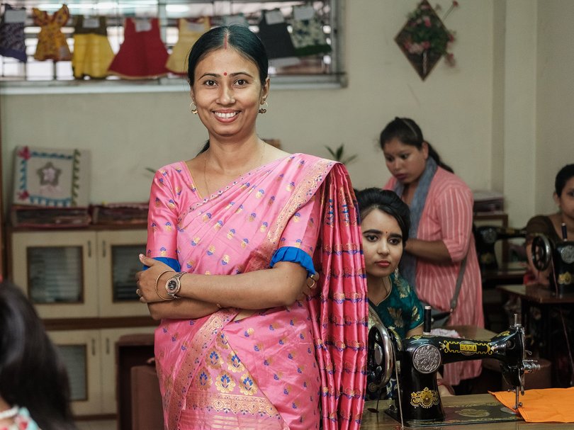 Lächelnde Frau im pinkfarbenen Sari in einer geschäftigen Nähwerkstatt, umgeben von anderen Frauen an Maschinen.