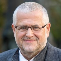 Porträt eines lächelnden Mannes mittleren Alters mit kurzen grauen Haaren, grauem Bart und Brille, trägt eine graue Jacke.