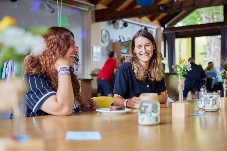 Zwei fröhliche Frauen sitzen an einem Holztisch; eine lacht ausgelassen, die andere lächelt in die Kamera.