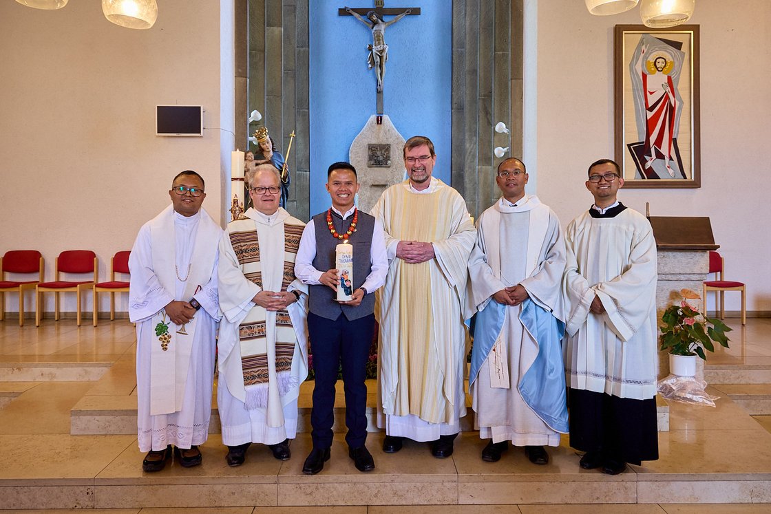 Sechs lächelnde Männer in geistlichen Gewändern posieren in einer Kirche. Einer hält eine verzierte Kerze. Kruzifix und Kunst im Hintergrund.
